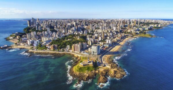 A foto mostra uma vista aérea de uma cidade litorânea com uma extensa faixa de prédios altos ao fundo e várias praias ao longo da costa. Em primeiro plano, há uma pequena península com um farol histórico e construções ao redor, cercada por águas azuis e áreas rochosas. O mar é amplo e ocupa grande parte da imagem, com tons que variam do verde próximo à costa ao azul profundo mais distante. O céu está claro, indicando um dia ensolarado. (futuro)