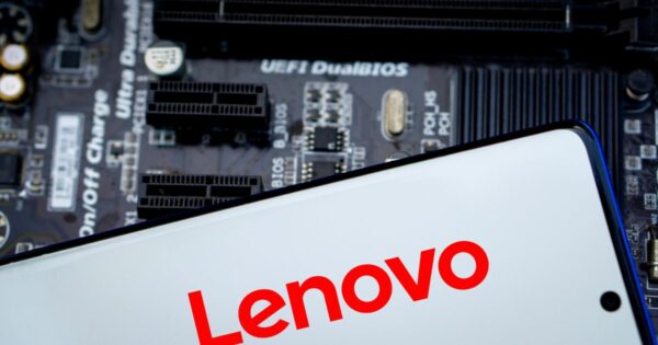 A foto mostra um dispositivo eletrônico com a marca Lenovo em destaque, escrita em letras vermelhas sobre fundo branco, provavelmente na tela de um smartphone ou tablet. Ao fundo, desfocado, há uma placa-mãe com componentes visíveis, incluindo slots de expansão e texto técnico como “UEFI DualBIOS”. A imagem transmite um contexto tecnológico, associando a marca Lenovo a hardware e sistemas. Infraestrutura
