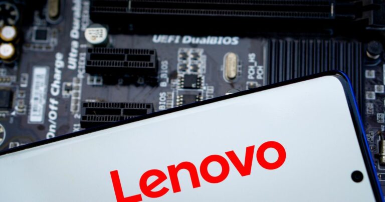 A foto mostra um dispositivo eletrônico com a marca Lenovo em destaque, escrita em letras vermelhas sobre fundo branco, provavelmente na tela de um smartphone ou tablet. Ao fundo, desfocado, há uma placa-mãe com componentes visíveis, incluindo slots de expansão e texto técnico como “UEFI DualBIOS”. A imagem transmite um contexto tecnológico, associando a marca Lenovo a hardware e sistemas. Infraestrutura