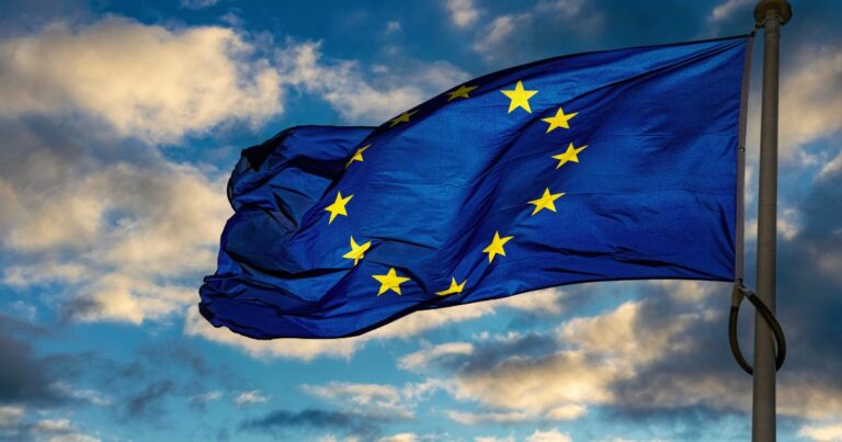 A imagem mostra uma bandeira da União Europeia hasteada em um mastro, ondulando ao vento. O fundo é composto por um céu parcialmente nublado, com nuvens em tons claros contrastando com áreas de azul intenso, sugerindo um cenário ao entardecer ou amanhecer. A bandeira é azul com doze estrelas douradas dispostas em círculo, símbolo clássico da UE que representa unidade e harmonia entre os países membros. A iluminação natural destaca o brilho das estrelas e o movimento do tecido, transmitindo dinamismo e imponência. (flexibiliza)