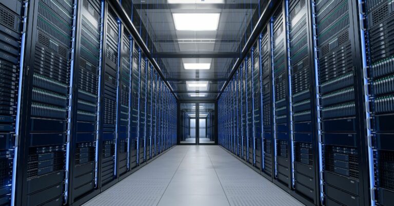 A foto mostra o interior de um corredor de data center, com fileiras de racks de servidores alinhadas dos dois lados. Cada rack contém múltiplos equipamentos eletrônicos organizados verticalmente, com luzes indicadoras verdes e azuis acesas. O ambiente é iluminado por painéis de luz no teto, refletindo no piso claro e criando uma aparência limpa e moderna. A perspectiva central dá profundidade à imagem, destacando a simetria e a infraestrutura tecnológica. (IEA), computação, legrand, para representar futuro data center da Huge Networks