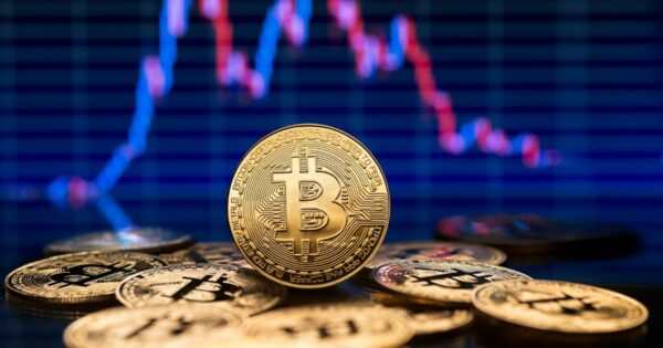 A foto mostra várias moedas douradas com o símbolo do Bitcoin em destaque. Uma moeda está posicionada verticalmente no centro, exibindo claramente o logotipo “₿” e detalhes gravados ao redor. Ao fundo, desfocado, há um gráfico financeiro com linhas vermelha e azul sobre um fundo escuro, sugerindo variação de preços ou análise de mercado. A imagem transmite um contexto relacionado a criptomoedas e finanças digitais.