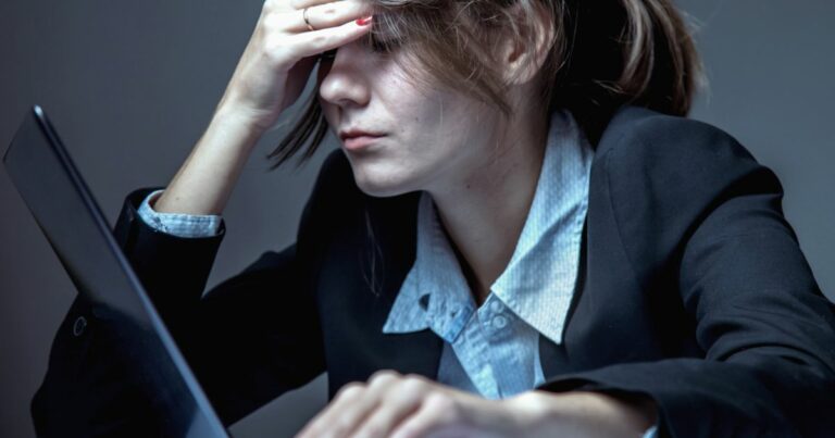 A imagem mostra uma pessoa vestindo um traje formal, incluindo um blazer preto e uma camisa social clara com colarinho. A pessoa está sentada diante de um laptop aberto, com uma das mãos apoiada na cabeça e a outra sobre o teclado, sugerindo que está envolvida em uma atividade de trabalho ou estudo. O ambiente parece interno, com iluminação suave e um fundo neutro, sem elementos adicionais visíveis. A postura inclinada e o foco no computador indicam concentração ou esforço mental. (crime)