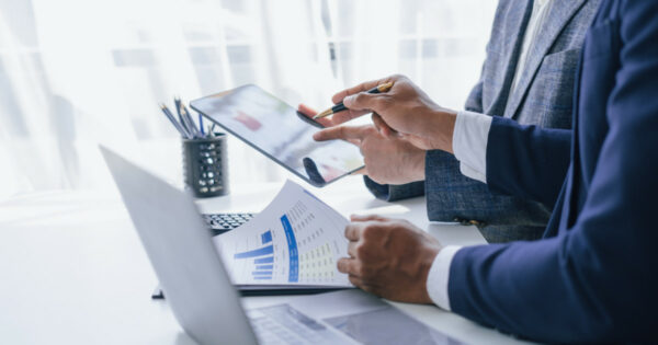 Duas pessoas em ambiente de escritório analisando gráficos impressos e dados em um tablet, com laptops sobre a mesa, representando uma situação de consultoria