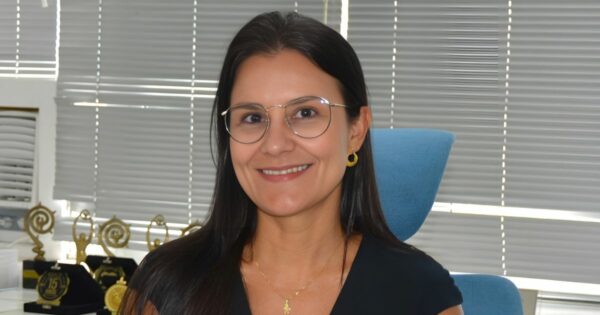 A imagem mostra uma pessoa sentada em uma cadeira de escritório azul, usando uma blusa preta e acessórios dourados — incluindo brincos circulares e um colar delicado. Ao fundo, há uma parede com persianas cinza fechadas e uma mesa onde estão dispostos vários troféus dourados, sugerindo um ambiente corporativo ou uma sala de premiações. A iluminação é neutra e destaca os elementos do cenário. (justiça)
