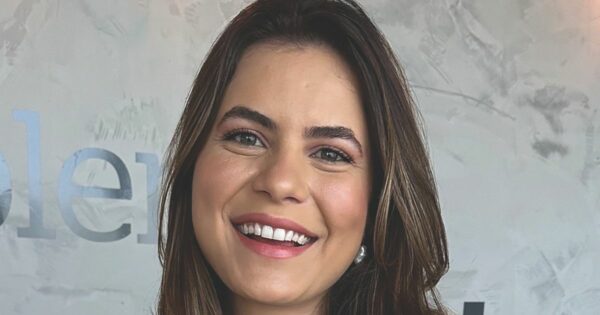 A imagem mostra uma pessoa de cabelos castanhos com mechas mais claras, soltos e levemente ondulados, posicionada em frente a uma parede texturizada em tons de cinza. Ao fundo, é possível ver parte de um texto aplicado à parede, com letras grandes e metálicas parcialmente visíveis. A iluminação suave destaca tanto a textura do fundo quanto os fios de cabelo, criando um ambiente moderno e minimalista. (IA)