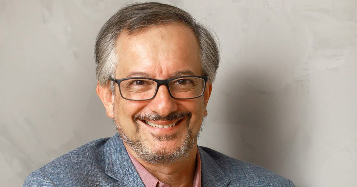 A imagem mostra a parte superior de um homem posicionado diante de uma parede de textura clara. Ele veste um blazer azul acinzentado sobre uma camisa social rosa, compondo um visual formal e discreto. O enquadramento destaca a roupa e os ombros, com iluminação suave que cria um tom neutro e profissional. (Nelson Campelo, CEO da Semantix)