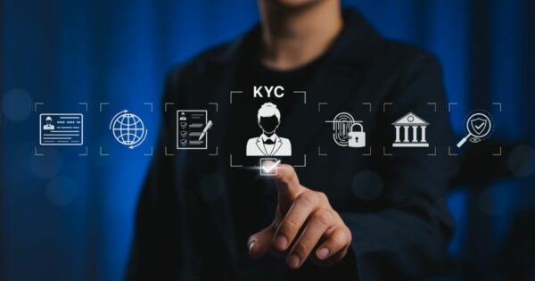 A imagem mostra uma pessoa em segundo plano, vestindo um traje escuro, com o rosto desfocado para manter anonimato. Em primeiro plano, há uma interface gráfica com ícones relacionados a processos de verificação e segurança digital. No centro, destaca-se um ícone com a silhueta de uma pessoa e a sigla “KYC” (Know Your Customer), indicando procedimentos de identificação de clientes. Ao redor, há outros ícones: um documento de identidade, um globo representando conectividade global, uma lista com marcações, um cadeado com impressão digital, um edifício que remete a instituições financeiras e um escudo com um símbolo de verificação. A pessoa está tocando um quadrado iluminado abaixo do ícone central, sugerindo interação com um sistema digital. O fundo é azul escuro com iluminação suave, transmitindo um ambiente tecnológico e corporativo.