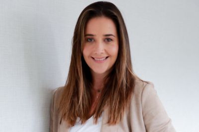 A pessoa está vestindo um blazer bege sobre uma blusa branca, posicionada diante de uma parede clara. A composição é simples e elegante, típica de retratos profissionais.