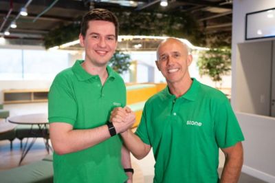 Duas pessoas de camisa verde da Stone Duas pessoas vestem camisas polo verdes com logo da empresa “Stone”. Elas estão em um aperto de mãos amistoso dentro de um escritório amplo, com móveis modernos e plantas ao fundo.