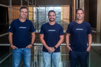 Três pessoas com camisetas da Softplan Três pessoas posam lado a lado, todas usando camisetas azul-marinho com o logotipo “Softplan”. Elas estão em um ambiente moderno com estruturas metálicas e vidro.