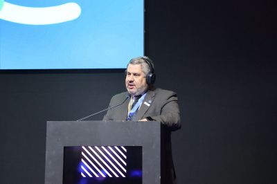 A pessoa está em um palco ou auditório, posicionada atrás de um púlpito preto com detalhes iluminados em linhas diagonais. Usa fones de ouvido e está vestida com terno escuro e gravata azul. Ao fundo, há uma tela com parte de um logotipo ou gráfico em azul e branco, sugerindo um evento corporativo ou conferência.