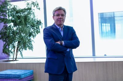 A pessoa está em pé, com braços cruzados, vestindo um terno azul escuro e camisa clara. O ambiente é interno, com janelas grandes ao fundo, luz natural entrando e uma planta decorativa próxima, transmitindo um cenário corporativo.