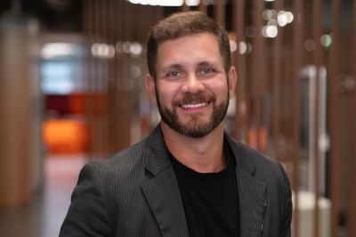 Pessoa de blazer escuro em corredor corporativo Pessoa vestindo blazer risca‑de‑giz escuro e camiseta preta. O fundo é um corredor moderno com divisórias de madeira e iluminação quente.