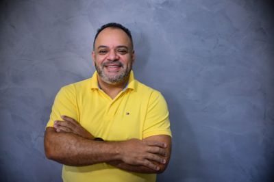 Pessoa com braços cruzados, vestindo camisa polo amarela, posicionada diante de um fundo texturizado em tom azulado, transmitindo postura confiante e informal.