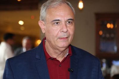 A pessoa está vestindo um blazer azul escuro sobre uma camisa social vermelha, com um microfone preso à roupa. O fundo é interno, com iluminação quente e elementos desfocados, sugerindo um ambiente de evento ou reunião.