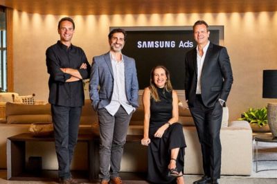 Grupo de quatro pessoas em frente a tela “Samsung Ads” Quatro pessoas em trajes sociais posam em um ambiente elegante com sofá e iluminação aconchegante. Ao fundo, há uma tela com o nome “Samsung Ads”.