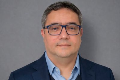Pessoa usando blazer escuro e camisa clara, em enquadramento frontal, diante de um fundo neutro em tom acinzentado, com aparência formal e corporativa.