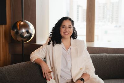 A pessoa está sentada em um sofá cinza, vestindo um conjunto elegante de blazer e calça claros, com camisa branca por baixo. Ao fundo, há uma janela ampla mostrando prédios urbanos e uma luminária metálica esférica ao lado, sugerindo um ambiente moderno e sofisticado.