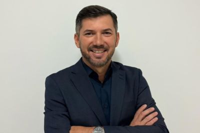 Pessoa vestindo blazer escuro com braços cruzados, posicionada diante de uma parede clara, transmitindo postura profissional e segura.