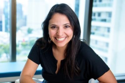 A pessoa está sentada em um ambiente corporativo, vestindo uma blusa preta simples. Ao fundo, há janelas amplas com vista para prédios urbanos, sugerindo um escritório moderno.