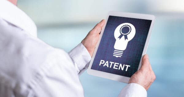 Pessoa segurando um tablet na horizontal, que exibe a imagem de uma lâmpada estilizada com um selo de certificação no centro e a palavra “PATENTE” em destaque abaixo. O fundo do dispositivo tem textura azul escura com linhas sutis, enquanto o ambiente ao redor é claro e desfocado, sugerindo um escritório ou espaço corporativo.