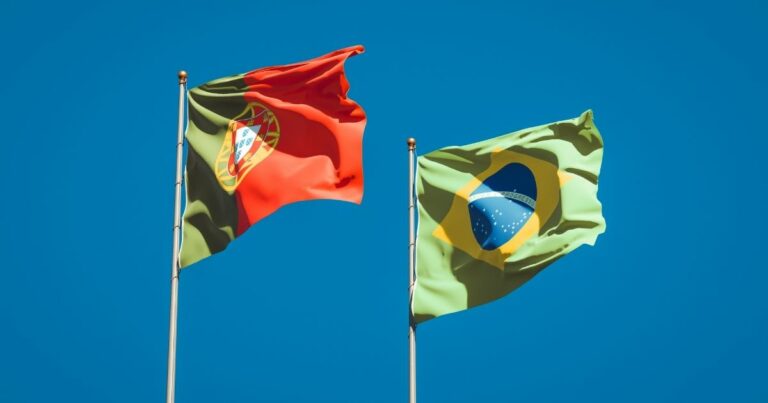 A imagem mostra duas bandeiras hasteadas contra um céu azul claro, criando uma composição vibrante e simbólica. Detalhes principais: Bandeira à esquerda: É a bandeira de Portugal, com as cores verde e vermelho divididas verticalmente. No centro, está o brasão nacional com o escudo e as esferas armilares, elementos históricos que representam a identidade portuguesa. Bandeira à direita: É a bandeira do Brasil, com fundo verde, um losango amarelo e um círculo azul contendo estrelas e a faixa branca com o lema “Ordem e Progresso”. Ambiente: Céu limpo e azul intenso, sem nuvens, destacando as cores das bandeiras. As bandeiras estão hasteadas em mastros metálicos, sugerindo um evento oficial, encontro diplomático ou celebração cultural. Essa imagem simboliza conexão histórica e cultural entre Brasil e Portugal, além de possíveis temas como cooperação internacional, laços linguísticos e parcerias estratégicas.