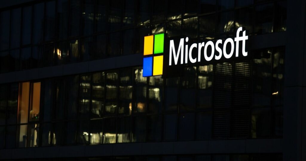A imagem mostra a fachada de um edifício corporativo com o logotipo da Microsoft iluminado em destaque. Detalhes principais: Texto visível: “Microsoft” escrito em brancoacompanhado do ícone característico da empresa — um quadrado dividido em quatro partes coloridas (vermelhoverdeazul e amarelo). Arquitetura: Estrutura moderna com grandes painéis de vidro refletindo luzes internas e externas. Iluminação noturnacom luzes acesas dentro do prédiocriando um contraste elegante com o fundo escuro. Ambiente: A cena sugere um horário após o expediente ou início da noitetransmitindo uma atmosfera corporativa e tecnológica. Essa imagem remete à inovaçãotecnologia e presença global da Microsoftreforçando sua identidade como líder em soluções digitais.