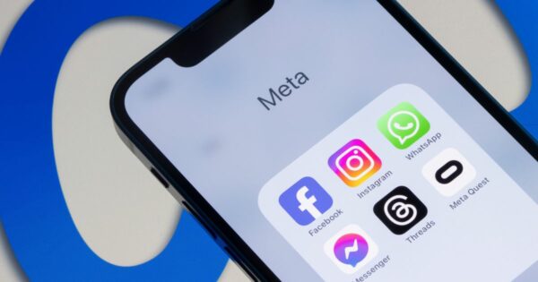 A imagem mostra um smartphone exibindo uma pasta com aplicativos da Meta, sobre um fundo com o logotipo da empresa em azul. Detalhes principais: Tela do dispositivo: Título da pasta: Meta Aplicativos visíveis: Facebook (ícone azul com “f”) Instagram (ícone colorido em degradê) WhatsApp (ícone verde com balão de conversa) Messenger (ícone roxo e azul) Threads (ícone preto com símbolo branco) Meta Quest (ícone branco com símbolo preto) Contexto visual: O fundo apresenta o logotipo da Meta em azul, parcialmente visível, reforçando a identidade da marca. Iluminação e cores: Tons claros e neutros na tela do smartphone contrastando com o azul vibrante do fundo, criando uma composição limpa e moderna. (Meta)