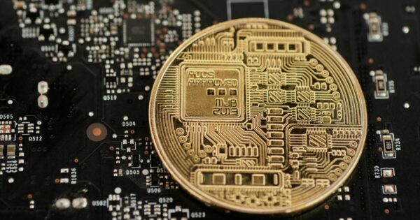 A imagem mostra uma moeda dourada com design inspirado em circuitos eletrônicos, posicionada sobre uma placa-mãe ou circuito eletrônico preto com diversos componentes e trilhas visíveis. O padrão da moeda inclui linhas e formas que remetem a tecnologia digital, sugerindo conceitos como criptomoeda, blockchain ou inovação tecnológica. A cena combina elementos físicos e digitais, transmitindo a ideia de finanças digitais e integração com tecnologia avançada. (Criptojud)