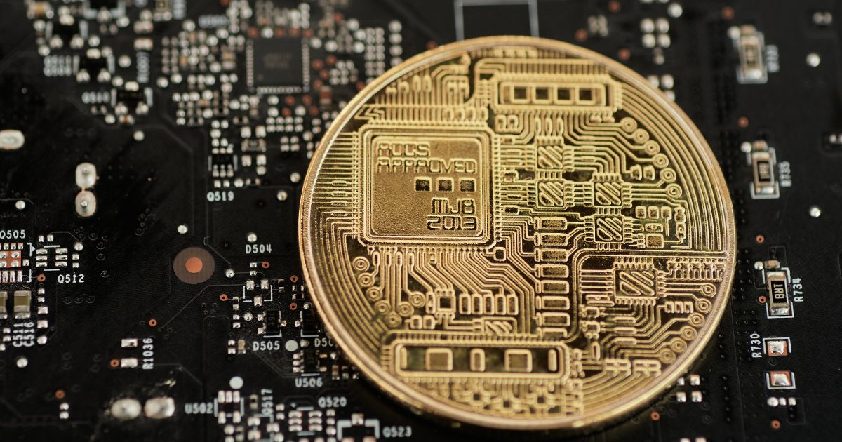 A imagem mostra uma moeda dourada com design inspirado em circuitos eletrônicos, posicionada sobre uma placa-mãe ou circuito eletrônico preto com diversos componentes e trilhas visíveis. O padrão da moeda inclui linhas e formas que remetem a tecnologia digital, sugerindo conceitos como criptomoeda, blockchain ou inovação tecnológica. A cena combina elementos físicos e digitais, transmitindo a ideia de finanças digitais e integração com tecnologia avançada. (Criptojud)
