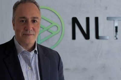 Pessoa vestindo blazer escuro e camisa listrada, posicionada diante de uma parede com logotipo corporativo