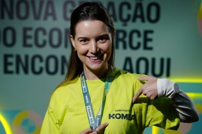 Uma pessoa usando camiseta amarela vibrante aponta para a estampa no peito, em um ambiente de evento com letreiro desfocado ao fundo.