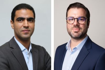 Duas pessoas usando trajes formais posam para foto em estúdio. A pessoa à esquerda veste um blazer escuro sobre camisa clara; a pessoa à direita usa um blazer azul-marinho com camisa clara.