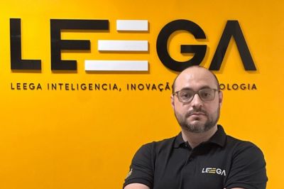 Pessoa com camisa polo preta da empresa "LEEGA", em pé diante de uma parede amarela com o logotipo da organização.