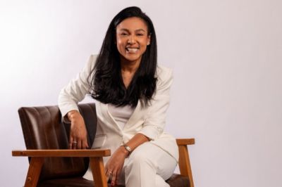 Uma pessoa usando conjunto branco elegante está sentada em uma cadeira de madeira, em pose sofisticada, diante de um fundo claro.