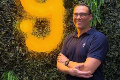 Uma pessoa vestindo camiseta polo azul-marinho está posicionada diante de uma parede com folhagens e um grande logotipo amarelo iluminado.
