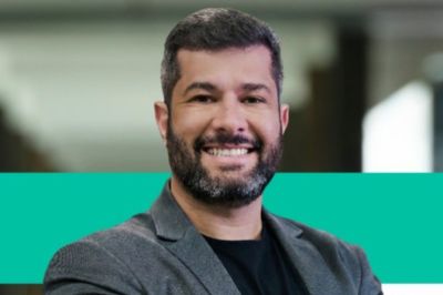 Pessoa vestindo blazer cinza e camisa preta, fotografada em ambiente corporativo com faixa verde no fundo.