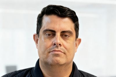 Uma pessoa com jaqueta escura aparece em foto profissional, com fundo claro e suave.