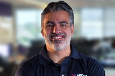 Uma pessoa usando uma camisa polo azul‑marinho aparece em um ambiente corporativo com mesas e janelas ao fundo, suavemente desfocados.