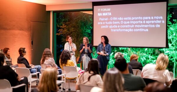 Um grupo de pessoas está reunido em uma sala para um painel de discussão. Três palestrantes sentam-se à frente do público, cada uma segurando um microfone enquanto conversam. Atrás delas, um telão exibe o título “IT Forum na Mata” e um texto sobre transformação contínua no RH. A sala tem grandes janelas que revelam vegetação iluminada do lado de fora. O público está sentado em fileiras de cadeiras brancas, atento ao debate.