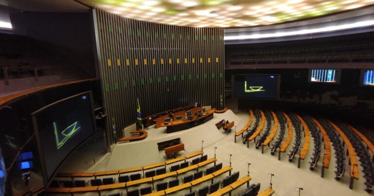 Vista ampla do plenário de uma casa legislativa, com fileiras semicirculares de assentos voltadas para uma mesa diretora central. Telões laterais exibem uma imagem gráfica em tons de verde. No fundo, há um grande painel vertical com detalhes metálicos e luzes nas cores verde e amarelo. O ambiente é iluminado por um teto amplo com pontos de luz distribuídos de maneira difusa. (Redata)