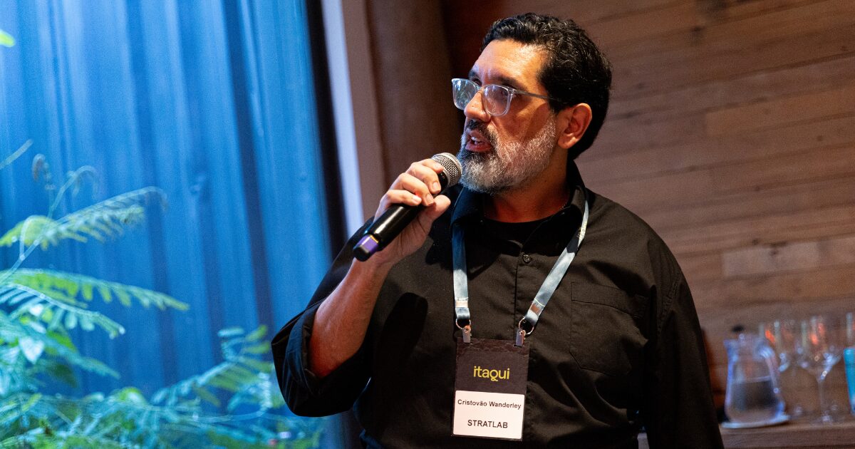 Pessoa segurando um microfone enquanto fala em um ambiente interno de evento. Ela veste uma camisa preta e um crachá pendurado no pescoço. Ao fundo, há uma parede de madeira, copos e jarros de vidro sobre uma mesa, além de um painel de vidro com plantas visíveis do lado de fora. (marketing)