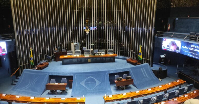 Vista interna ampla de um plenário legislativo em formato semicircular, com cadeiras distribuídas em fileiras curvas voltadas para a mesa principal. No centro, há uma grande área azul com o símbolo nacional estampado. A mesa diretora fica posicionada diante de um painel vertical composto por longas hastes metálicas. Telões laterais exibem transmissões da sessão, e bandeiras estão posicionadas ao lado da área central. O ambiente é iluminado de forma difusa, destacando o espaço amplo e formal. (Alcolumbre)