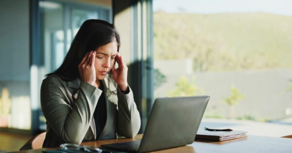 Uma pessoa sentada à mesa em um ambiente iluminado pela luz natural segura a cabeça com ambas as mãos, em postura que sugere preocupação ou cansaço. À sua frente, há um notebook aberto, um caderno e um par de óculos sobre a mesa. O fundo mostra janelas amplas com vista para uma área externa verde e ensolarada. (Saúde mental trabalho)