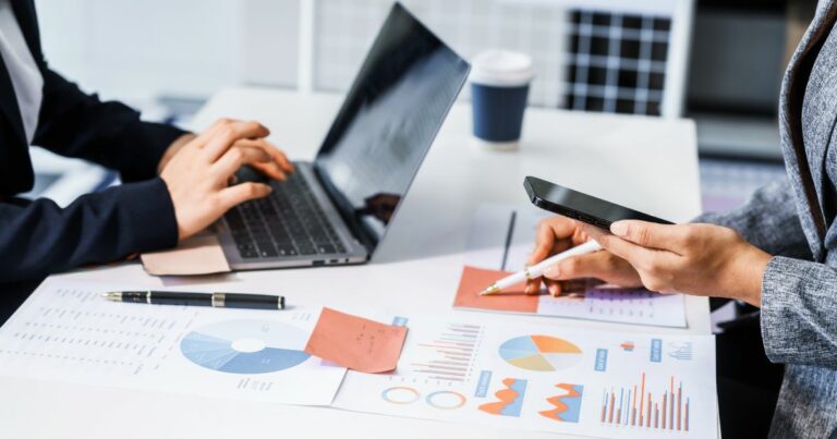 Duas pessoas trabalham juntas em uma mesa de escritório. Uma delas digita em um laptop, enquanto a outra segura um smartphone e utiliza uma caneta para apontar informações em gráficos impressos sobre a mesa. Os documentos exibem diagramas, gráficos de pizza, gráficos de barras e anotações coloridas, sugerindo análise de dados, planejamento financeiro ou revisão de relatórios. Também há canetas, papéis e uma xícara ao fundo, compondo um ambiente profissional de colaboração. (governança)