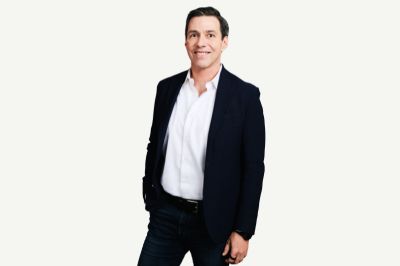 Uma pessoa em pose profissional veste uma camisa branca por baixo de um blazer azul-escuro. Ela está em pé, com uma das mãos no bolso e o corpo levemente inclinado para o lado, posicionada diante de um fundo claro e uniforme.