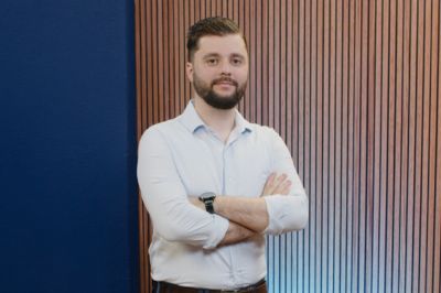 Uma pessoa com camisa social clara e mangas dobradas aparece com os braços cruzados diante de uma parede composta por painéis estreitos de madeira, ao lado de uma área azul.