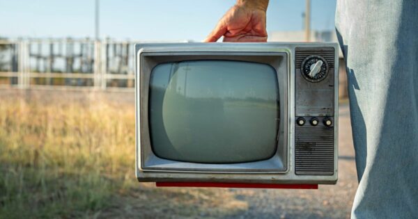 A imagem mostra uma televisão antiga sendo carregada por uma pessoa. A mão está segurando a alça superior do aparelho, que possui um design retrô, com bordas metálicas, tela curva e botões físicos na lateral direita — incluindo seletores circulares e pequenos botões alinhados horizontalmente. A televisão está sendo transportada ao ar livre, em um ambiente com vegetação seca e gramado amarelado ao fundo. Estruturas metálicas e postes aparecem desfocados ao longe, sugerindo uma área industrial ou rural. A pessoa que segura o aparelho veste uma roupa em tom azulado, visível apenas parcialmente na lateral direita da imagem. A iluminação é natural, com luz direta do sol, criando sombras suaves e realçando a textura desgastada da televisão analógica.