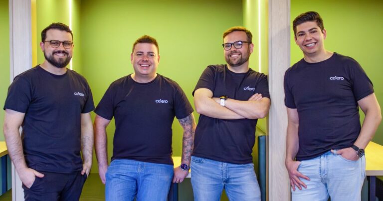 A imagem mostra quatro pessoas posicionadas lado a lado em um ambiente interno com paredes em tom de verde vibrante. Todas estão vestindo camisetas escuras com o logotipo “celero” estampado no lado esquerdo do peito. As duas pessoas à esquerda estão em pé, com os braços relaxados ao longo do corpo. A terceira pessoa está em pé com os braços cruzados. A pessoa à direita está encostada em uma coluna de madeira clara, com uma das mãos no bolso. O ambiente possui mesas de madeira clara ao fundo, criando um aspecto moderno e organizado. A iluminação é uniforme, destacando as cores das paredes e das roupas. (Celero)